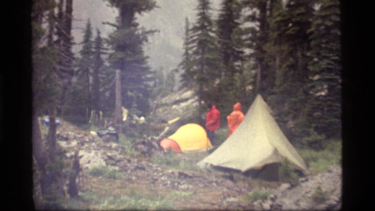 1977 8mm frame near Sapphire Lake, Montana, showing hikers in bright rain jackets beside yellow and beige tents in a forest campsite.