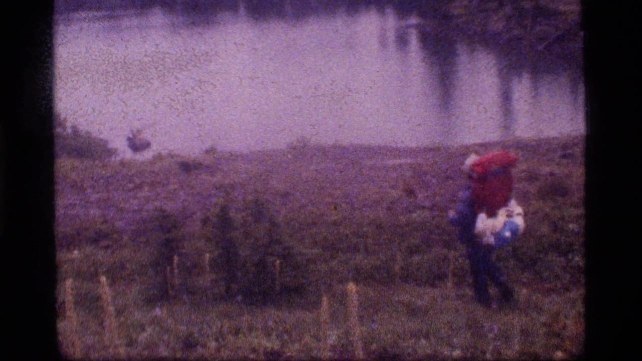 A backpacker in red gear stands near open, wet ground by water.