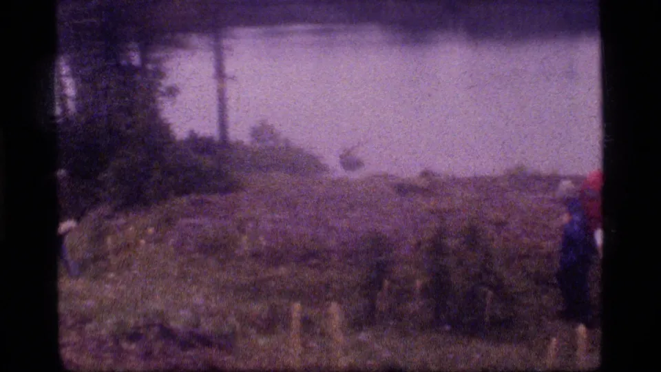 Soft 8mm frame from the Montana hiking clip showing damp open ground, water beyond, and a partially visible backpacker at frame edge.