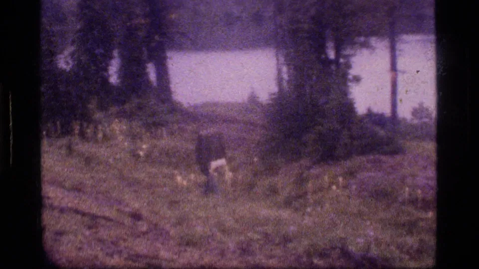 1977 8mm frame near Sapphire Lake, Montana, with a backpacker moving through forested ground beside a bright lake or wetland opening.