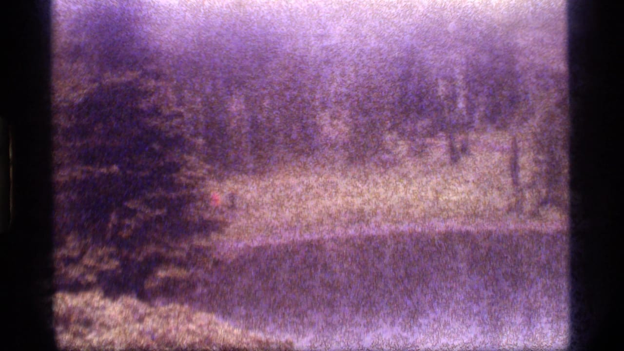 1977 8mm frame from Sapphire Lake, Montana, showing a distant red-shirted hiker near a pond and wooded bank.