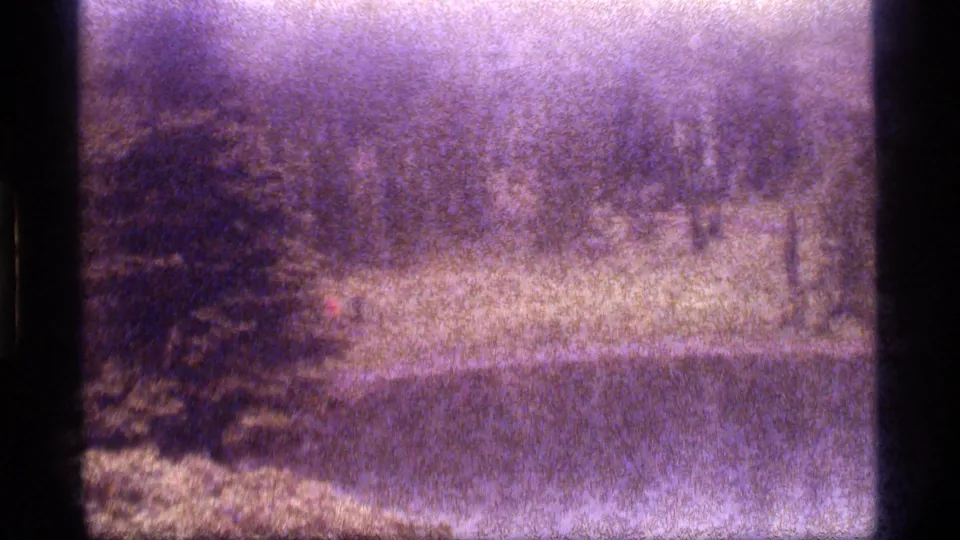 1977 8mm frame from Sapphire Lake, Montana, showing a distant red-shirted hiker near a pond and wooded bank.