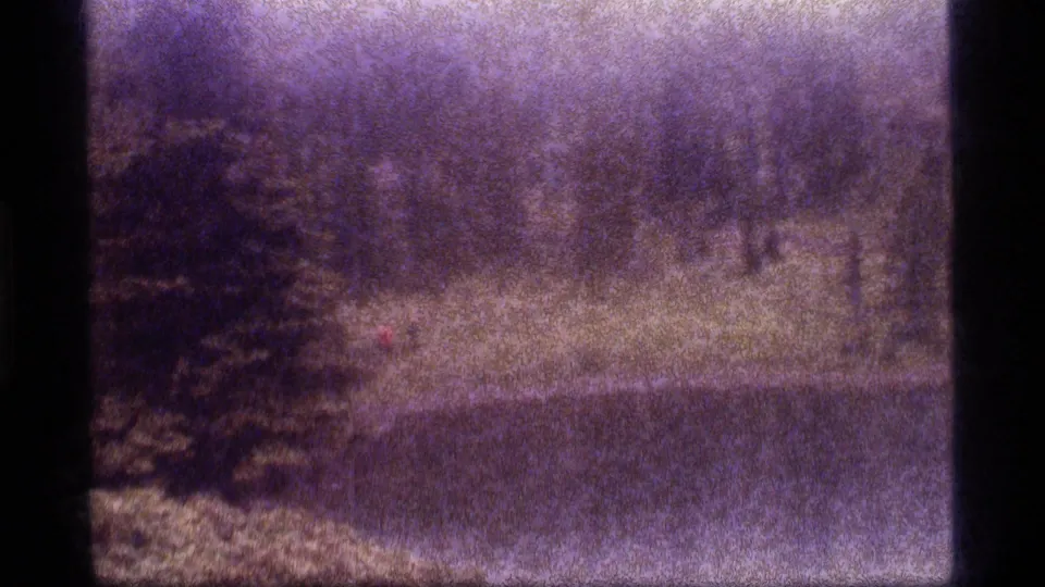 Soft 1977 home-movie frame showing a red-shirted hiker on a grassy slope near Sapphire Lake woodland water.