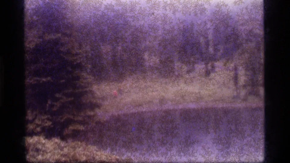 Purple-tinted 8mm frame from Sapphire Lake, Montana, with a distant hiker near a pond below dense trees.