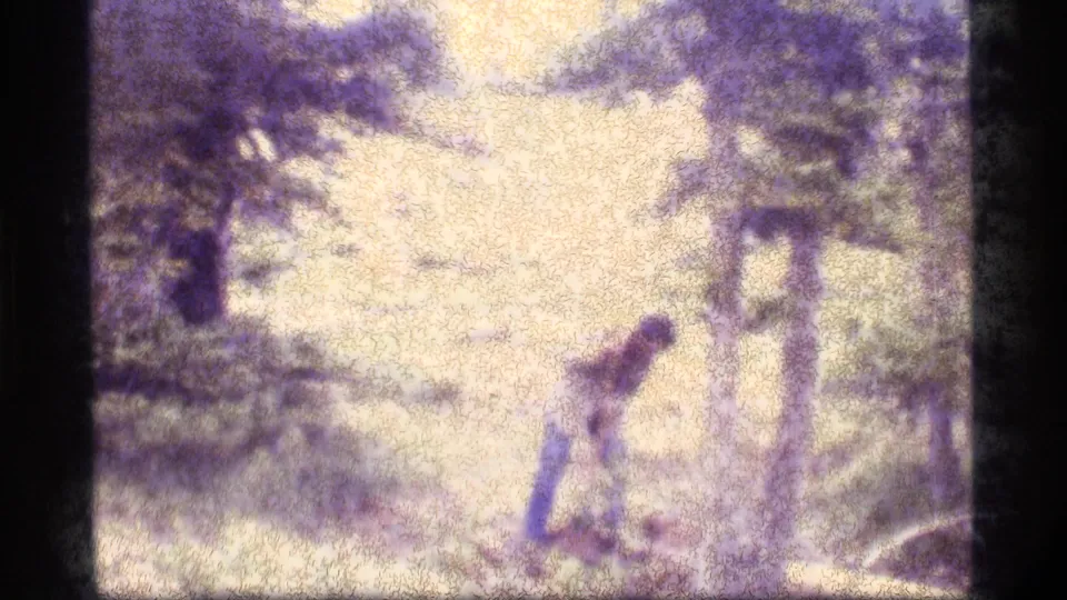 The man remains bent over near the ground in the Sapphire Lake woods, filmed from a handheld home-movie viewpoint in 1977.