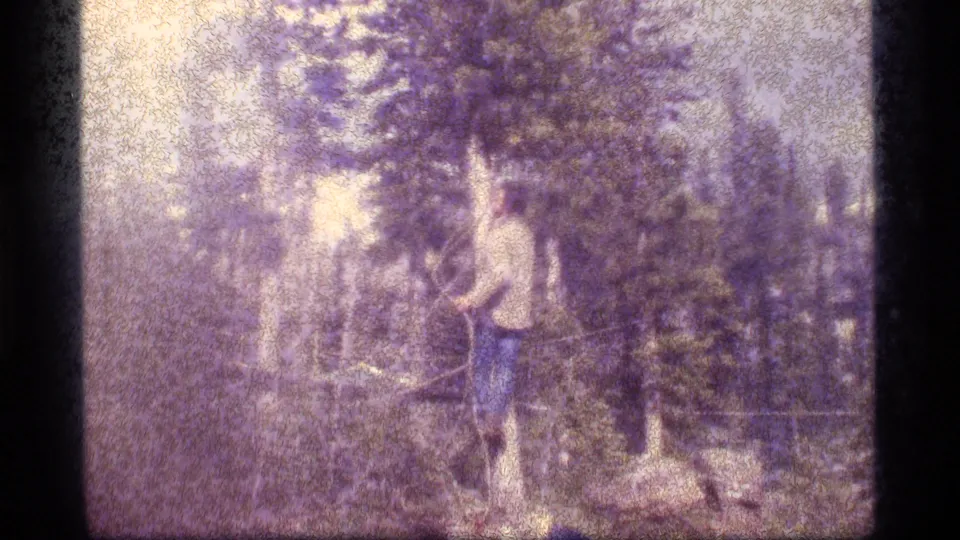 1977 Sapphire Lake, Montana frame of a person in a light shirt and dark pants standing in the forest, possibly working with a line or rope.