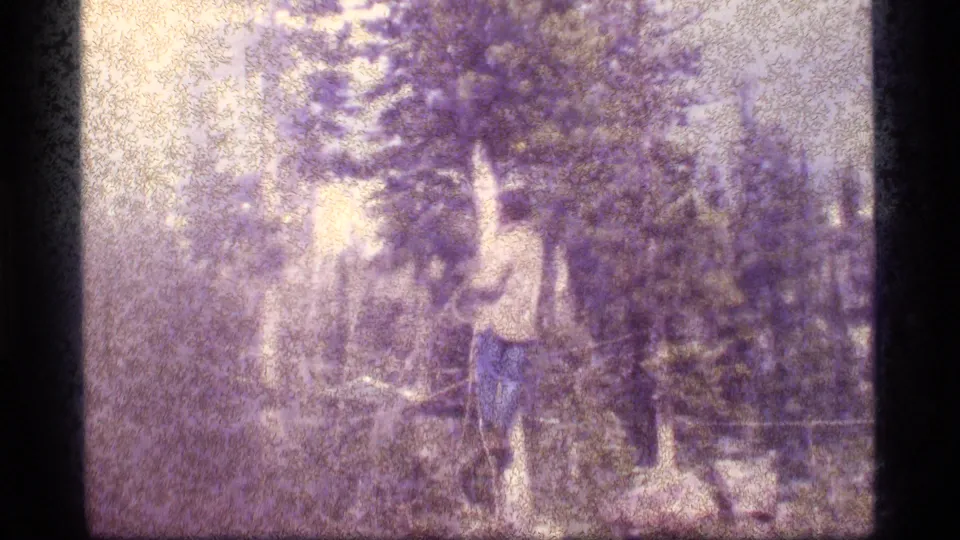 1977 8mm frame from Sapphire Lake, Montana, showing a man reaching toward a high branch with rope among tall trees.