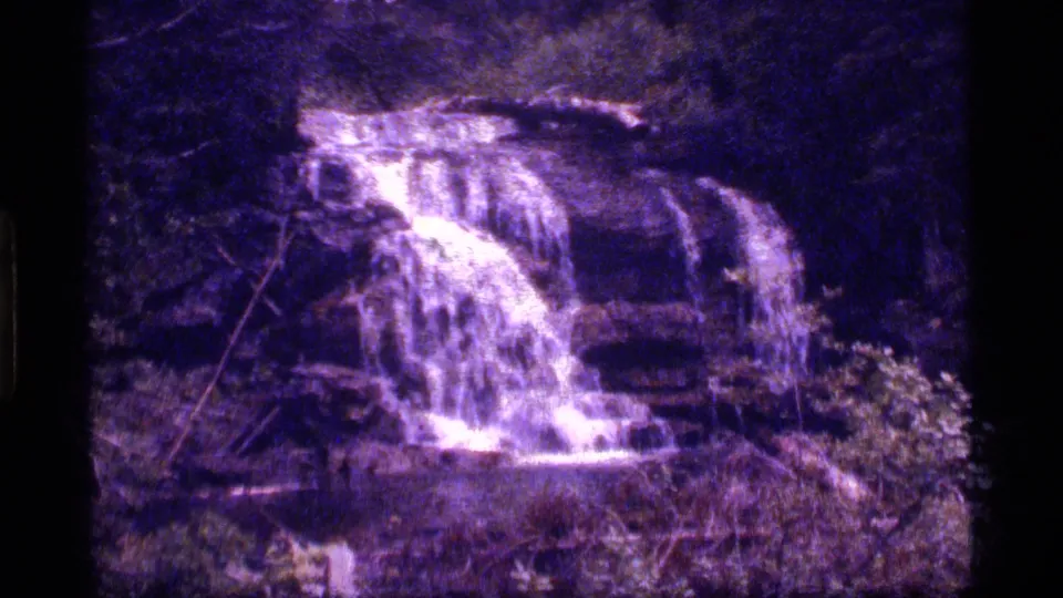 A small waterfall drops over rocks in the 1977 Sapphire Lake, Montana footage, framed in dark forest shade with purple-tinted film color.