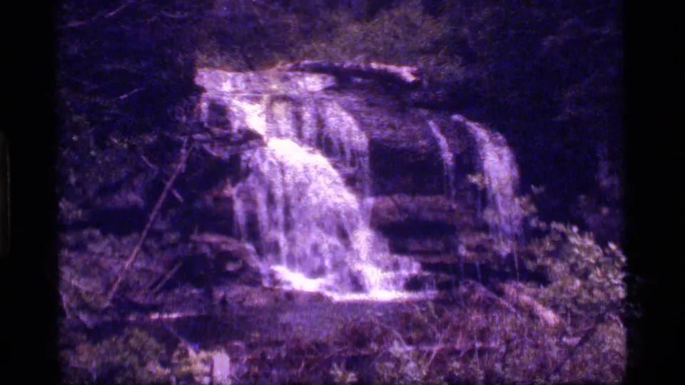 1977 8mm home-movie frame from Sapphire Lake, Montana, showing a small waterfall dropping over rock ledges amid dense trees.