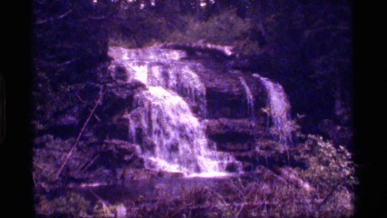 The waterfall remains centered in a purple-tinted 8mm view, with white water moving over rocks and dark forest around it.