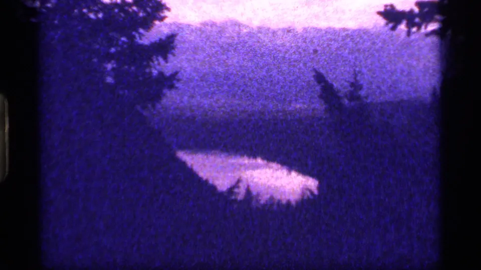 The Montana lake remains centered in a grainy 8mm frame, with dark trees around the edges and a soft hillside in the background.