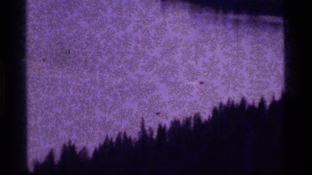 1977 8mm view of Sapphire Lake with a dark treeline silhouette above purple-shifted water and heavy film grain.