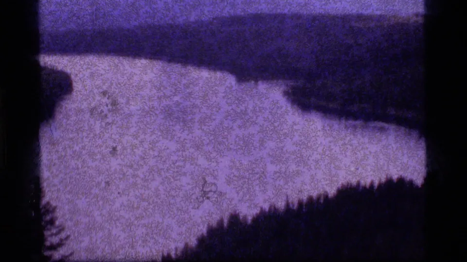1977 home-movie frame of Sapphire Lake with open water, wooded edges, and a dark shape near the shoreline area.