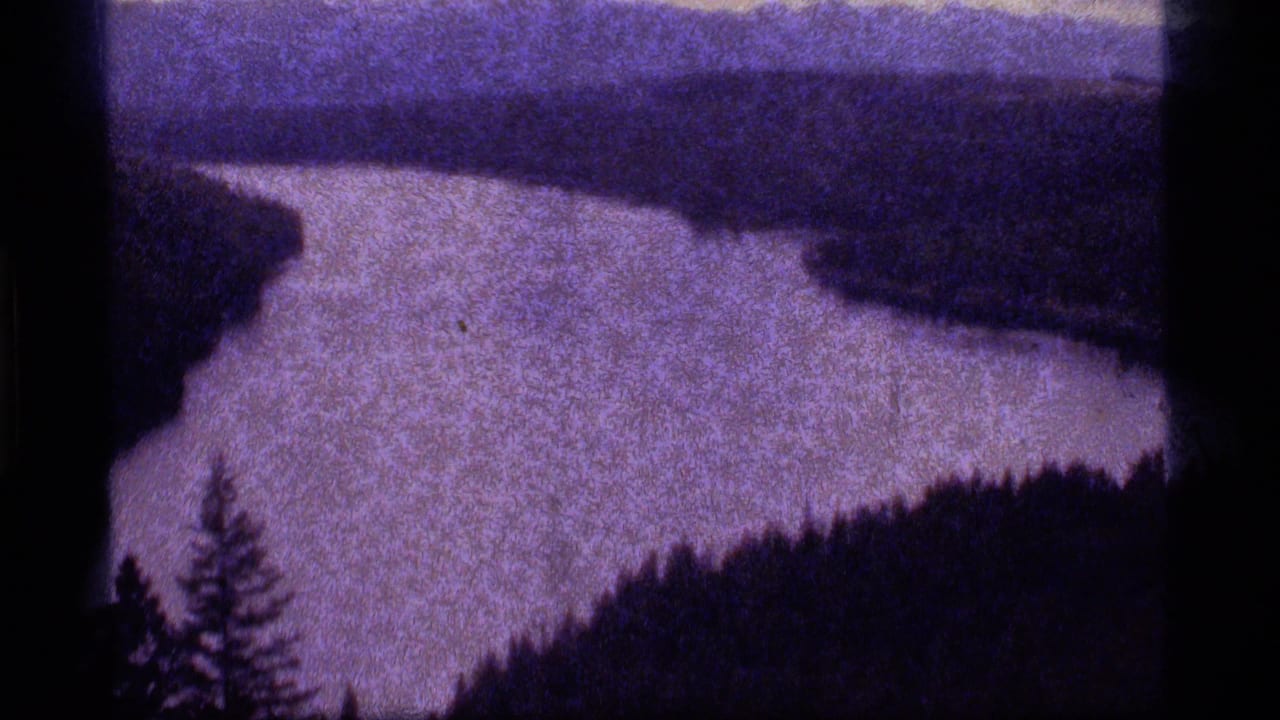 1977 8mm frame from Sapphire Lake, Montana, showing a purple-toned waterway between dark wooded banks.