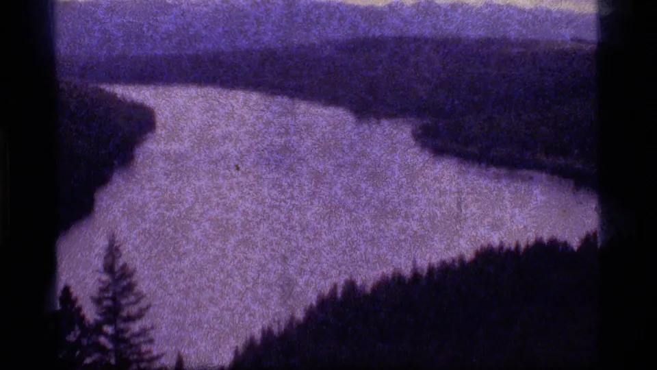 1977 8mm frame from Sapphire Lake, Montana, showing a purple-toned waterway between dark wooded banks.