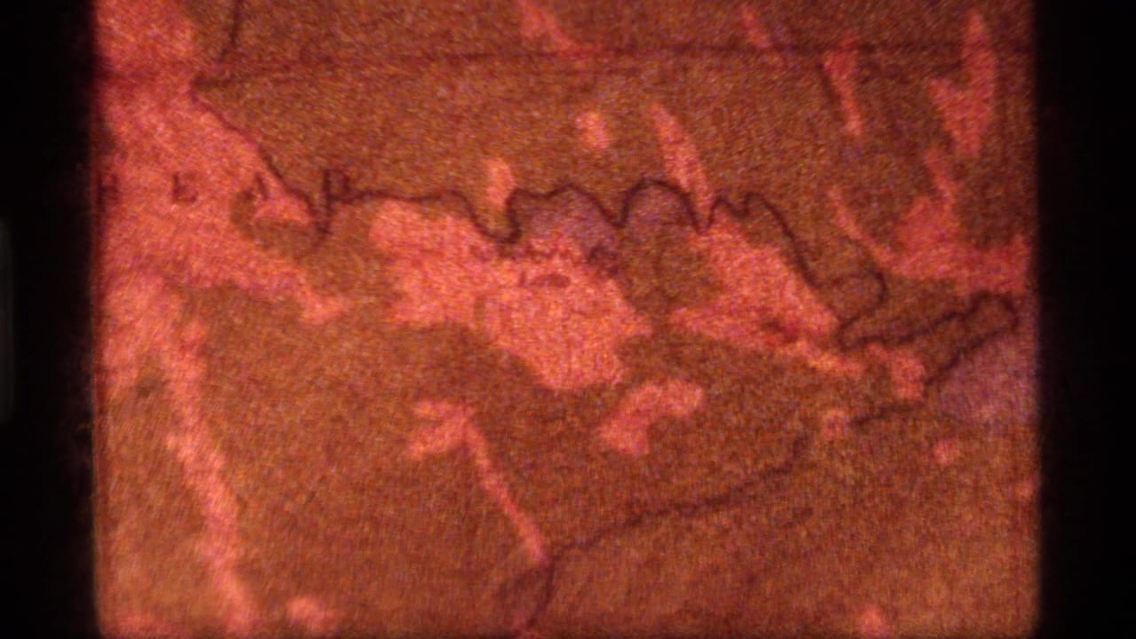 1977 Sapphire Lake, Montana home-movie frame showing a close view of a reddish map with analog grain and scratches.