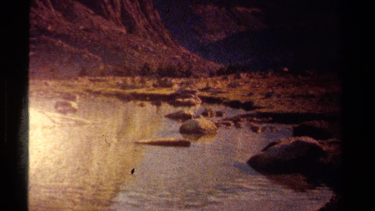1978 Lake Tahoe 8mm frame showing a dim rocky shoreline or stream below dark mountain slopes with warm light reflected on the water.