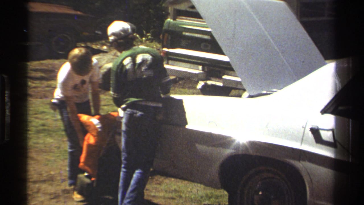 Adults stand near the open rear of a white sedan on grass in bright sun.
