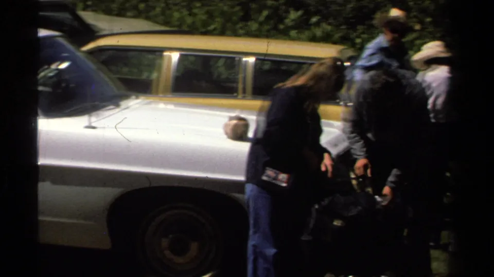 Adults sort through dark bags on grass beside a white car, with a yellow station wagon and foliage in the background.