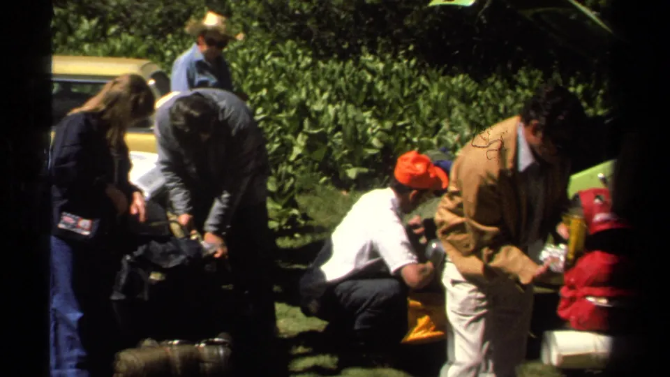 1978 Lake Tahoe roadside frame with adults handling bags and supplies near the car, shrubs, and bright grass.