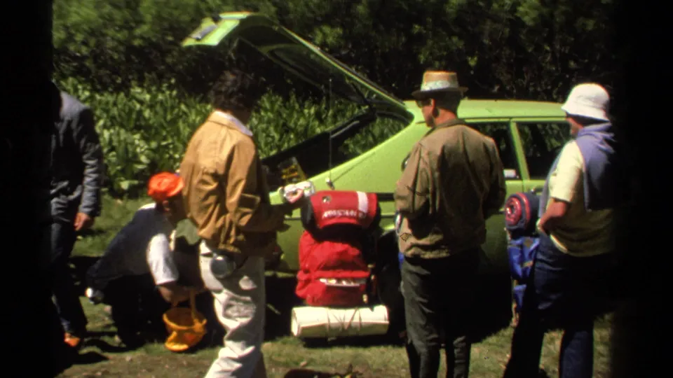 Lake Tahoe hikers in 1978 adjust or gather camping gear beside a green car, with trees and grass behind them.