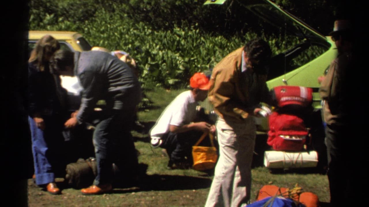 1978 Lake Tahoe home-movie frame showing adults sorting packs and supplies beside a bright green car on grass.