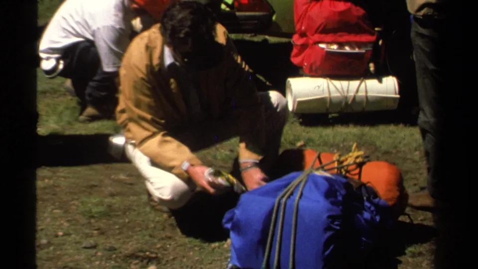 Close 1978 Lake Tahoe home-movie view of a man crouching to sort small supplies beside packed camping gear.
