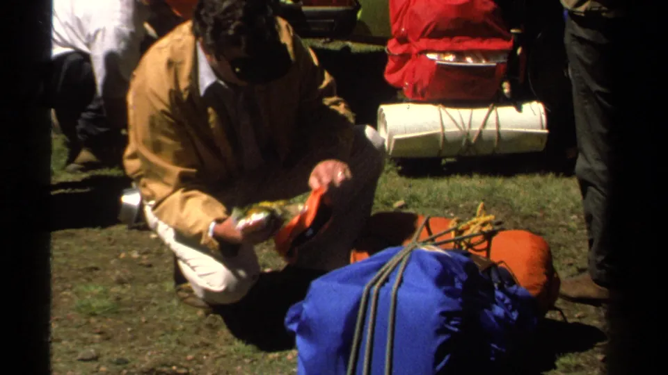1978 Lake Tahoe frame showing a man checking small supplies beside blue and red camping gear in bright sunlight.