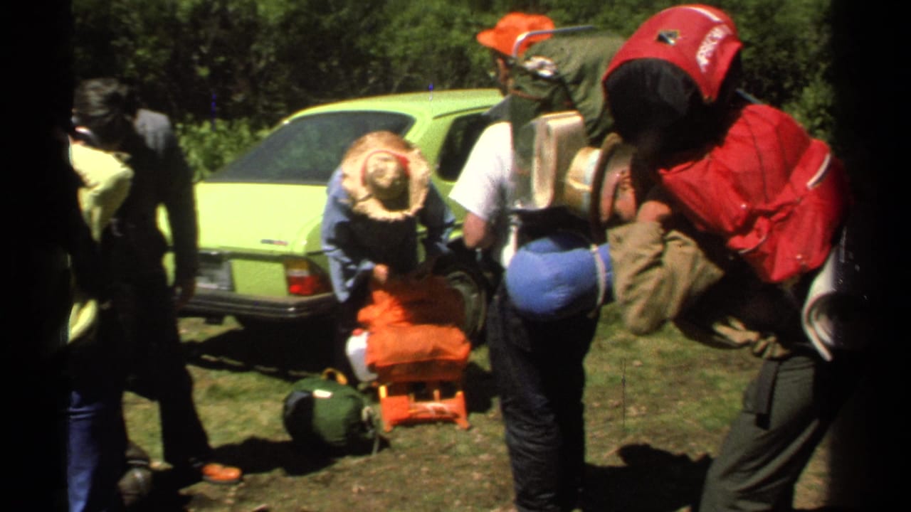 Hikers adjust backpacks and camping gear beside a lime-green car on grass.