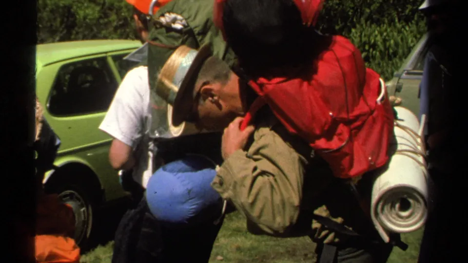 Close 1978 Lake Tahoe home-movie frame of hikers carrying large packs with a blue sleeping bag and rolled mat near a car.