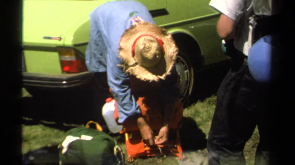 1978 Lake Tahoe frame with a straw-hatted man leaning over supplies near backpacks, a canteen, and the rear of a lime-green car.