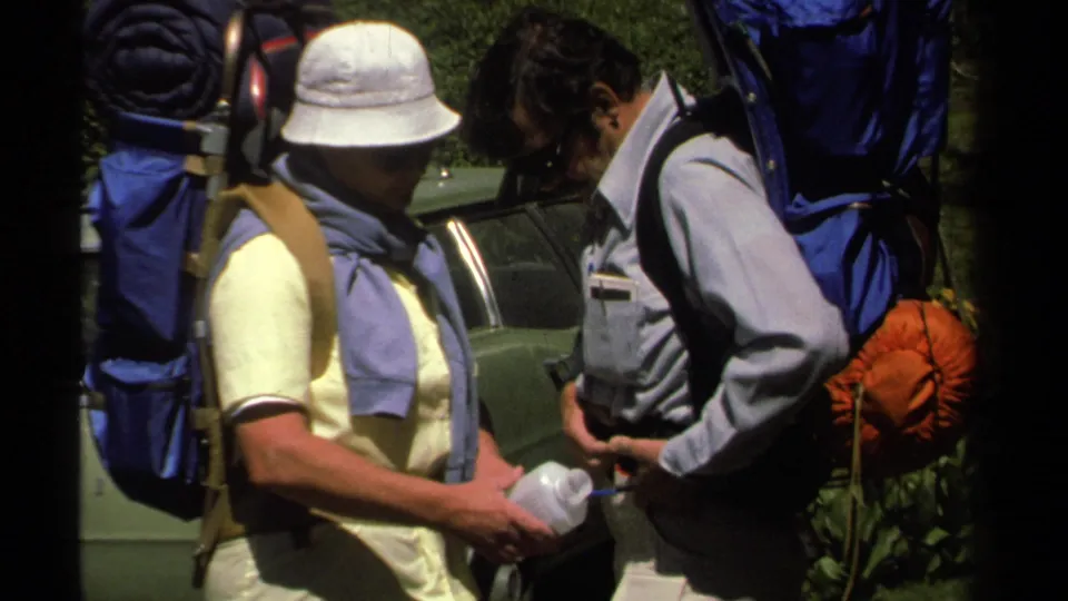 1978 Lake Tahoe home-movie frame showing two men with large backpacks checking a water container beside a parked car.