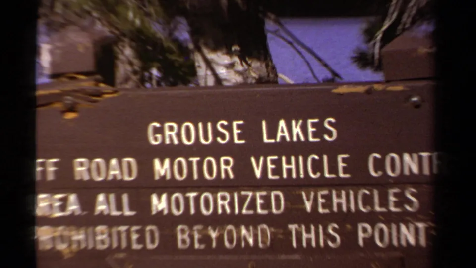 Close 1978 home-movie view of the Grouse Lakes control sign lettering, with tree branches and blue sky behind the board.