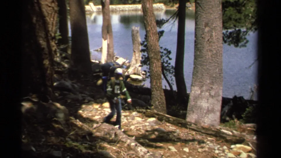 A backpacked hiker walks through a wooded lakeside area in the Lake Tahoe-region 1978 home-movie footage.