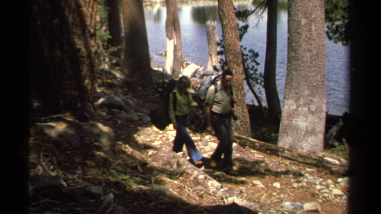 In 1978 Lake Tahoe footage, an older man and woman carry large packs along a sun-dappled rocky trail beside pine trees and lake water.