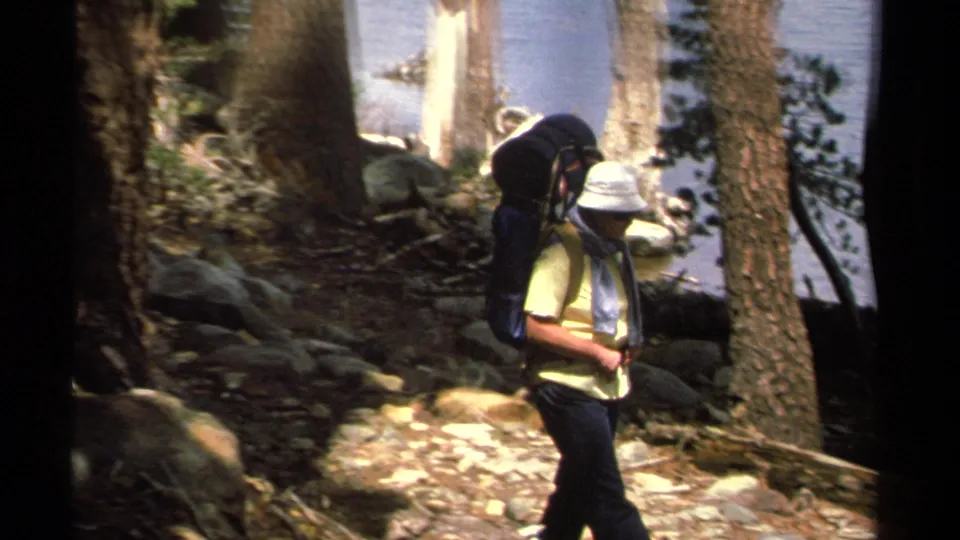A backpacker wearing a light hat passes through the Lake Tahoe forest trail area, with rocks, pine trunks, and blue water behind him.