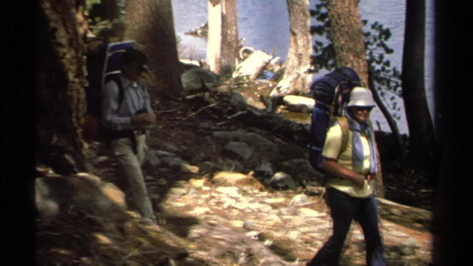 Two hikers with large backpacks move along a rocky wooded trail near Lake Tahoe in 1978 home-movie footage.