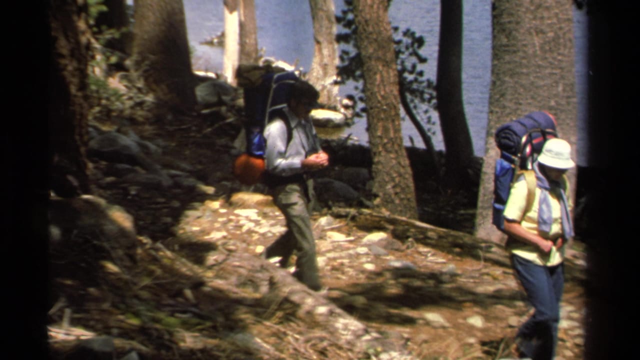 Two hikers with packs walk between trees with blue lake water behind them.