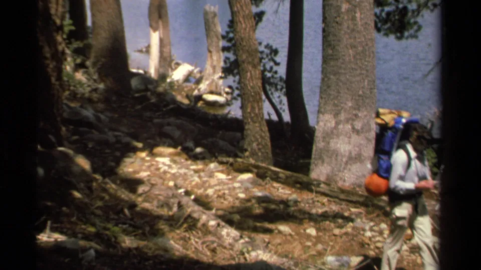 A backpacker with blue and orange gear leaves the frame on a shaded Lake Tahoe trail, with trees and water still visible.