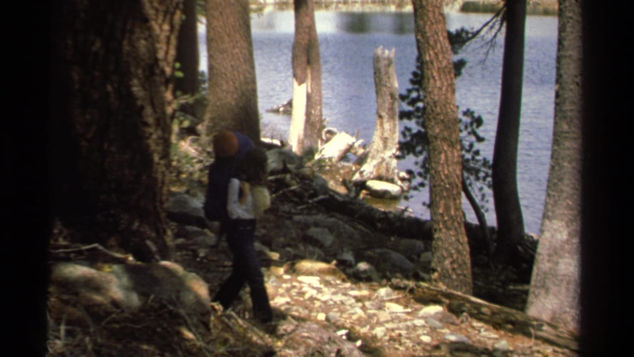 1978 Lake Tahoe home-movie frame showing a backpacker on a shaded pine forest trail with blue lake water behind.