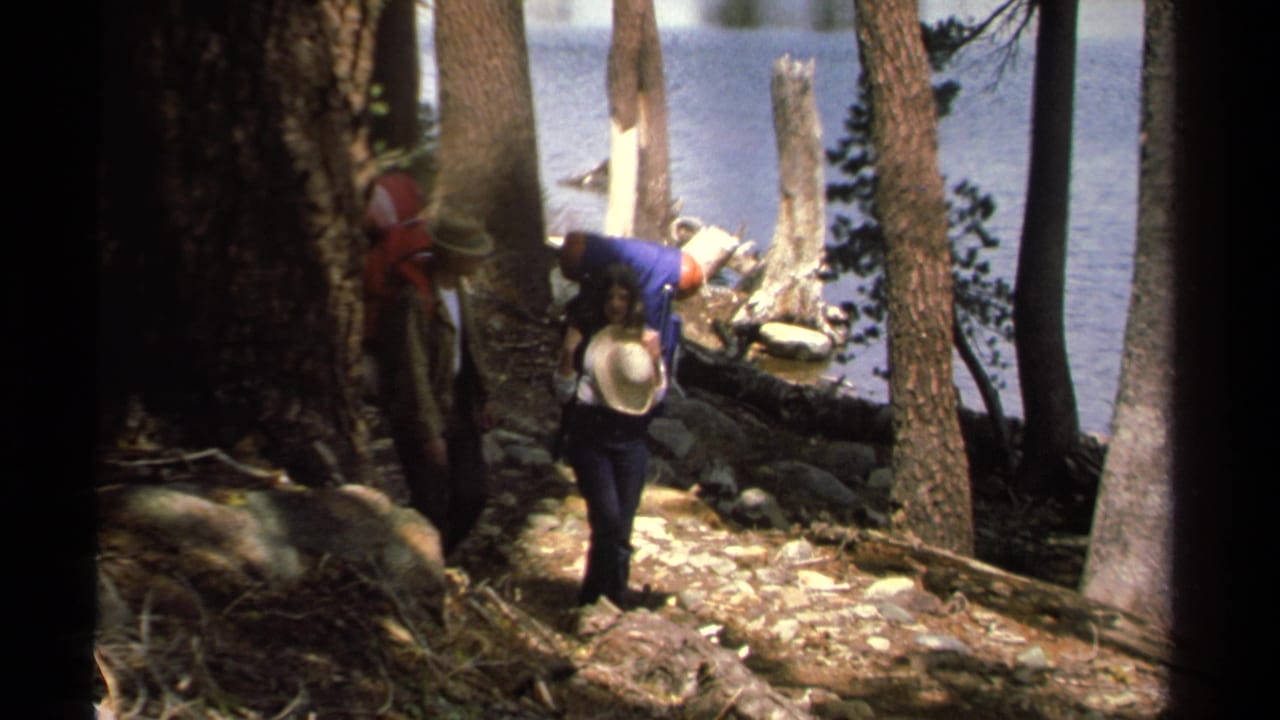 Two hikers with large backpacks walk near trees and lake water.
