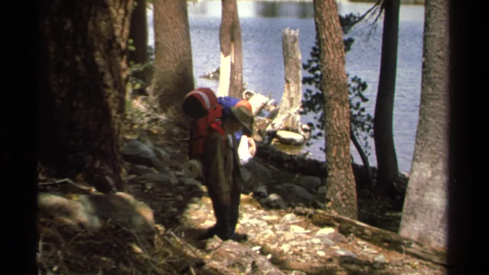 A 1978 Lake Tahoe trail frame with a hiker in a straw hat and red backpack following another person near lakeside trees.