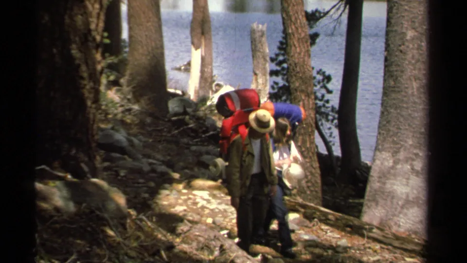 Two 1978 Lake Tahoe backpackers walk away along a rocky pine trail, with blue lake water visible beyond the trees.