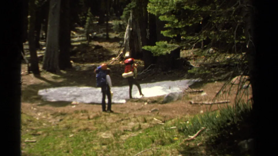 Two backpackers stand in a wooded Lake Tahoe area, one appearing to inspect terrain near rocks and open ground.