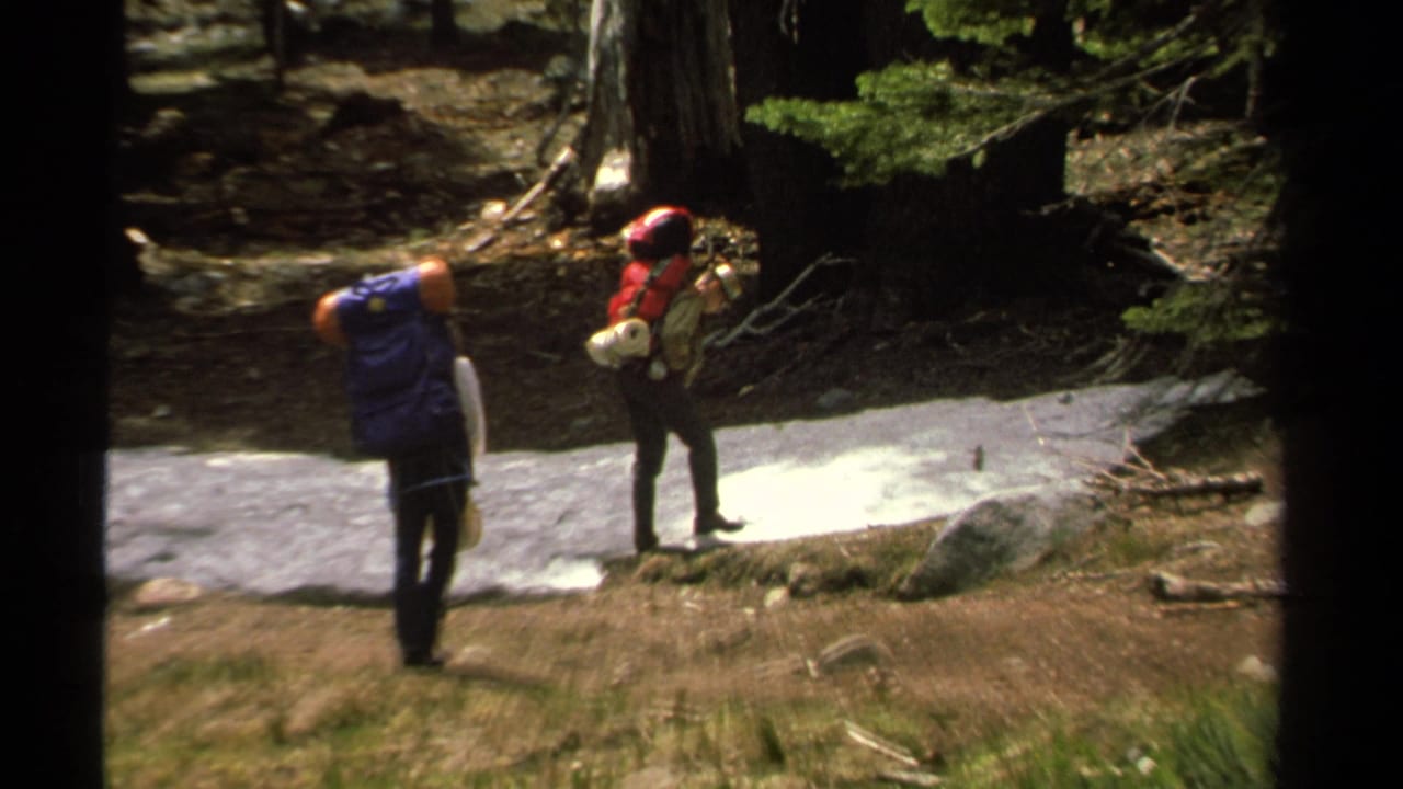 In 1978 Lake Tahoe footage, two hikers with red and blue backpacks stand near a lingering snow patch among trees.