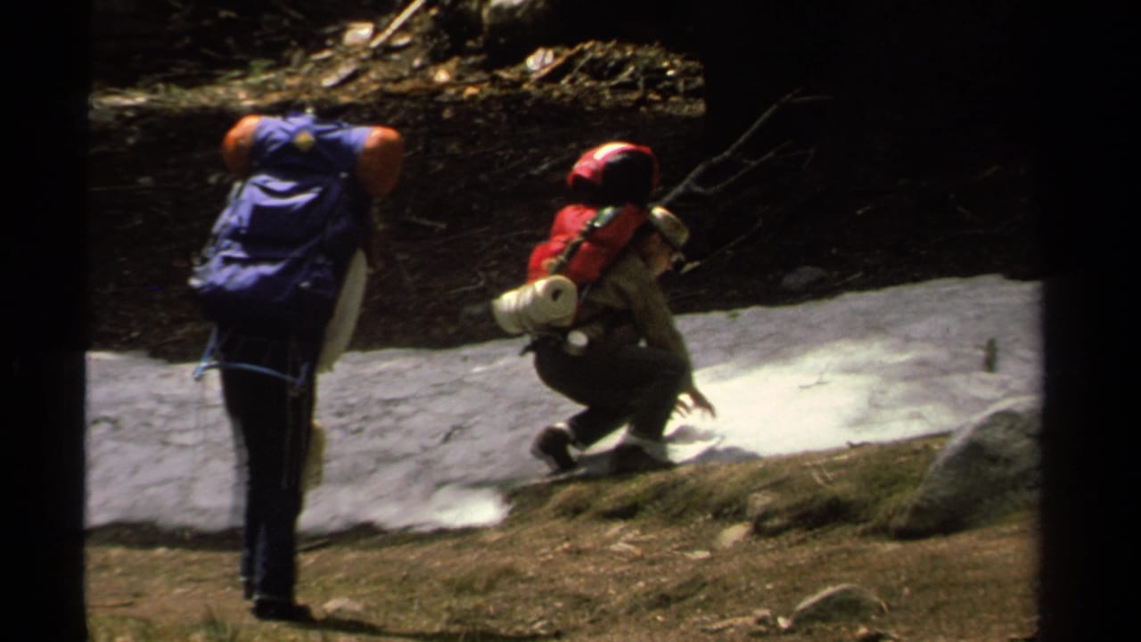 One hiker crouches beside snow while another stands nearby with a blue backpack.