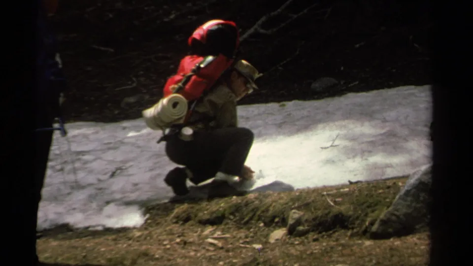 The red-backpack hiker crouches close to the remaining snow in the Lake Tahoe forest, partly framed by another person at left.