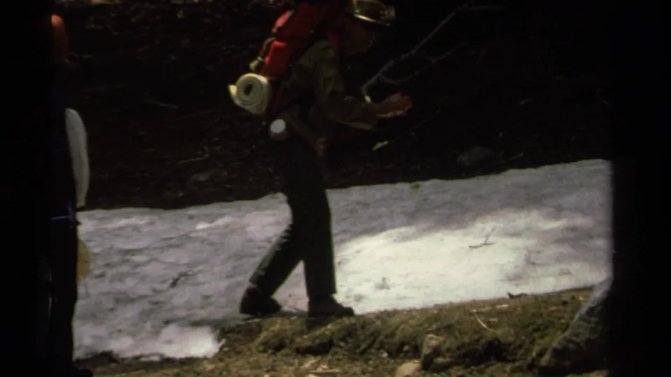 A hiker with a red backpack walks along the shaded Lake Tahoe forest trail beside a lingering white snow patch.