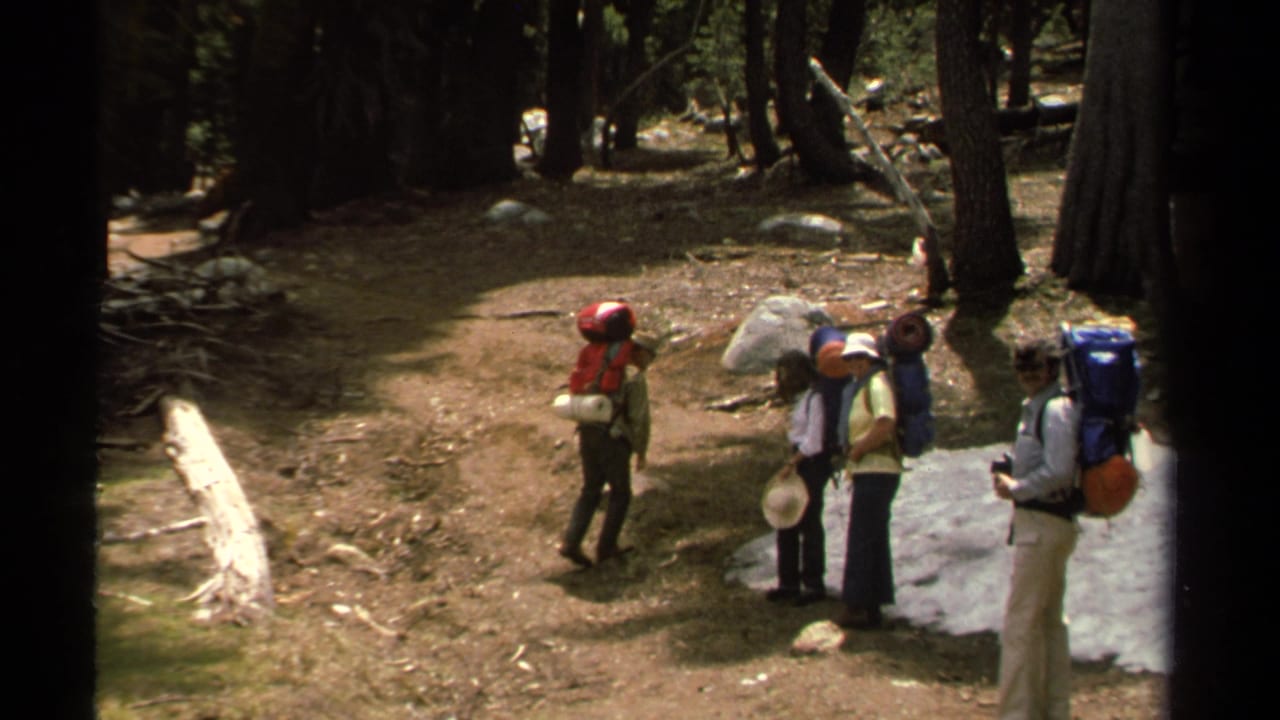 1978 Lake Tahoe home-movie frame showing four backpackers paused on a wooded trail with rocks, trees, and patches of snow.