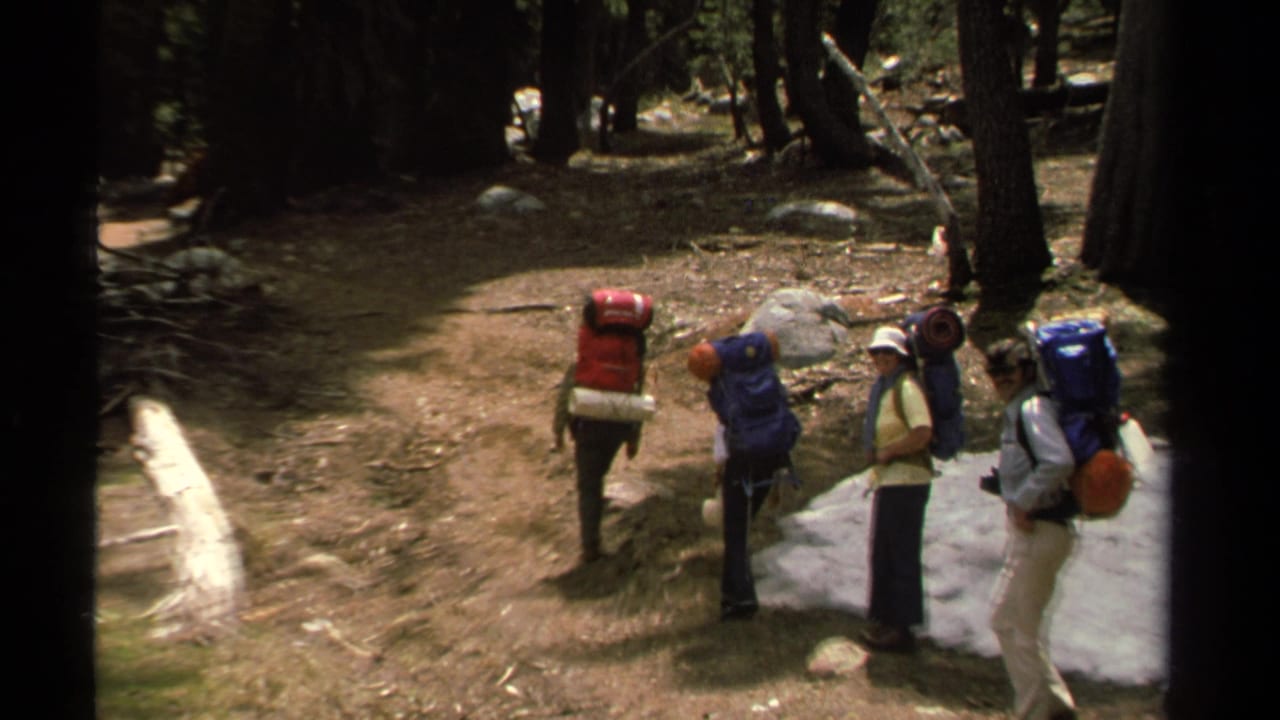 Backpackers begin moving along a dirt trail through trees beside a patch of snow.